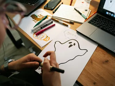 A person drawing Halloween-themed ghosts and pumpkins on paper at a desk with a  4k anime wallpaper