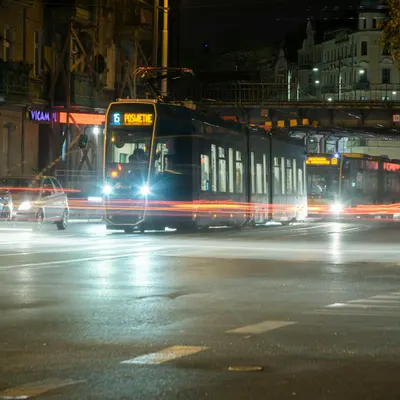 Tram moving at night with light trails in a bustling city street under a bridge. 4k city wallpaper