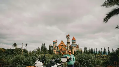 A person stands with a scooter before a castle-like structure surrounded by lush 4k fantasy wallpaper