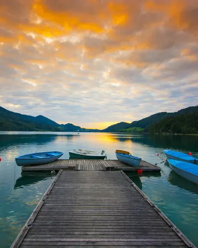 Idyllic sunset over a calm lake with moored blue boats, framed by tranquil mount 4k lake wallpaper