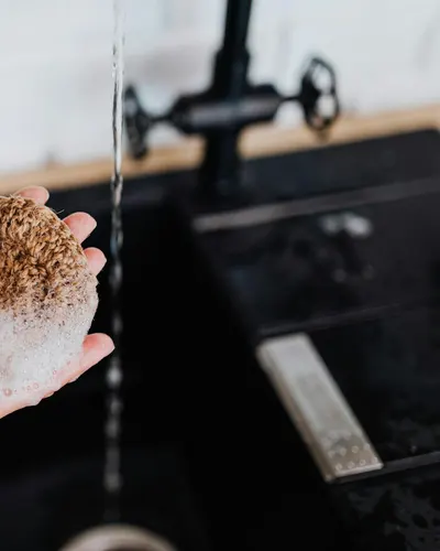 Close-up of a hand holding a natural sponge under running water at a sink. 4k minimal wallpaper