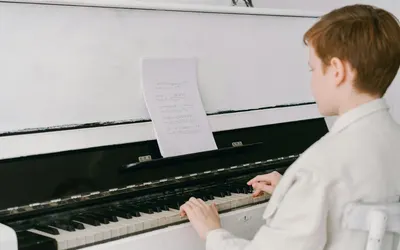 A young boy sits at an upright piano playing music indoors, focused on sheet mus 4k music wallpaper