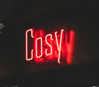 Glowing red neon 'Cosy' sign illuminating a dark urban street corner at night. 4k neon wallpaper