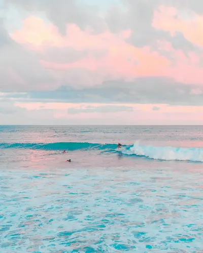 Surfer riding waves beneath a pastel sky at sunset. Tranquil ocean scene. 4k sunset wallpaper